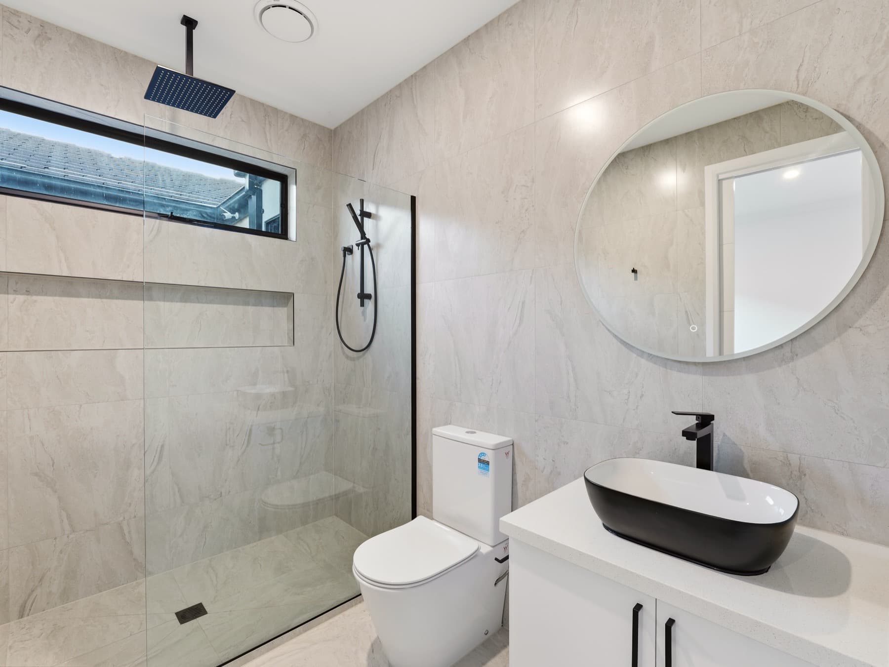 A bathroom with an open shower behind a black framed screen, round mirror over a black basin